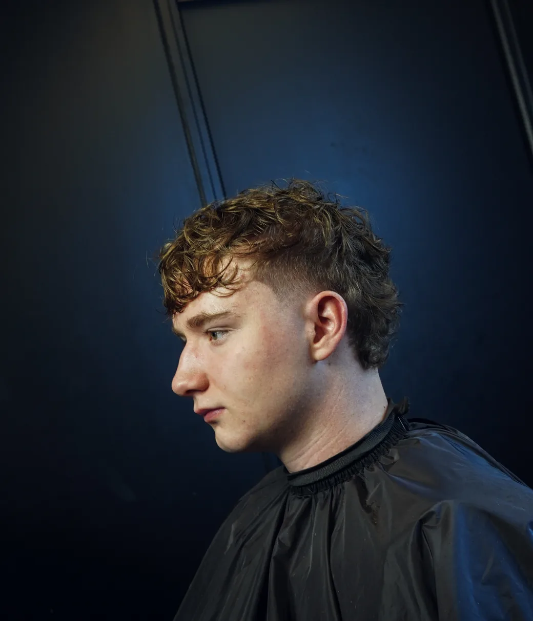 La photo de fin : un client roux de profil après la coupe, mulet bouclé, lumière rasante sur fond bleu nuit.